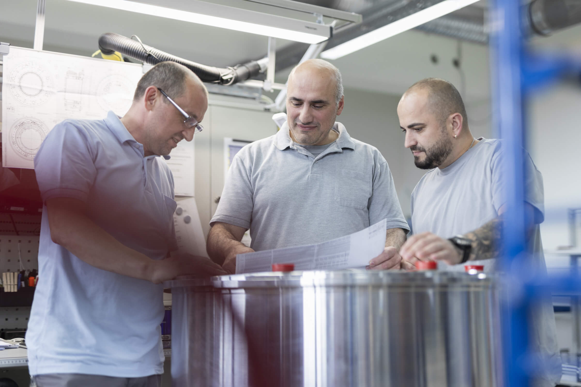Drei Techniker in einem Produktionslabor analysieren technische Zeichnungen über einem Metallzylinder.