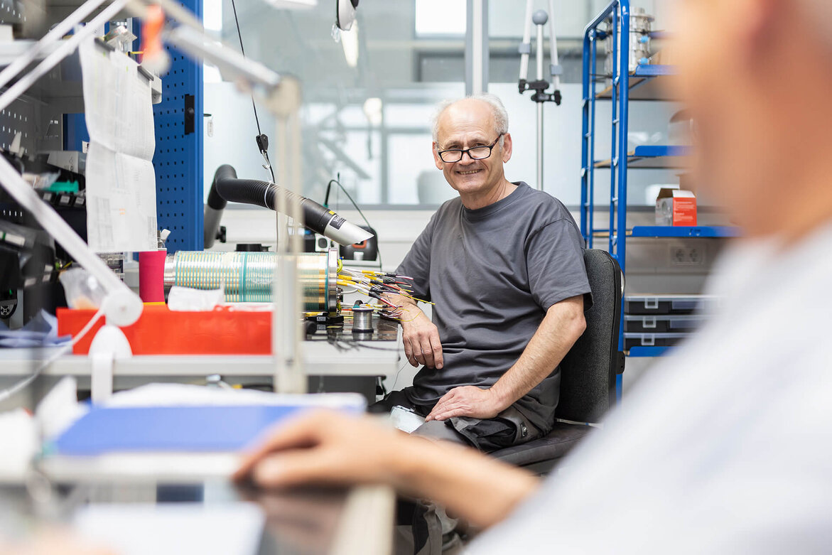 Älterer SCHLEIFRING Mitarbeiter sitzt in der Montage und lächelt.