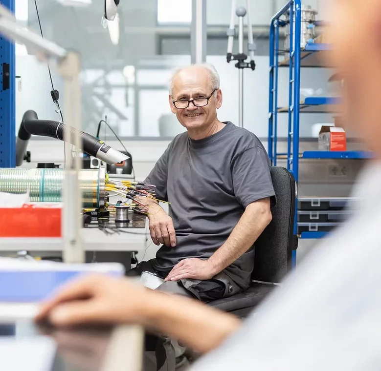 Mitarbeiter sitzt lächelnd an seinem Arbeitsplatz in der Elektronikfertigung.