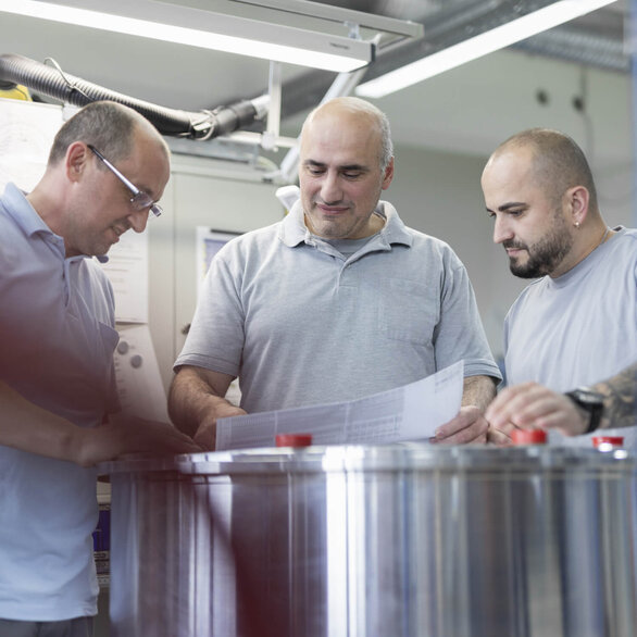 Drei Techniker in einem Produktionslabor analysieren technische Zeichnungen über einem Metallzylinder.